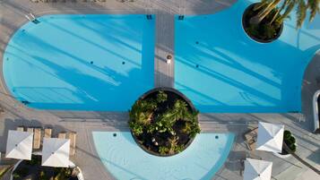An aerial view of a swimming pool with two separate sections, surrounded by lounge chairs and umbrellas, and a central landscaped area with plants.