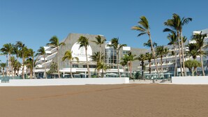 Un hotel moderno con un diseño arquitectónico único, que destaca una estructura circular y varios balcones. Palmeras bordean la playa, y el hotel está junto a una zona de arena.