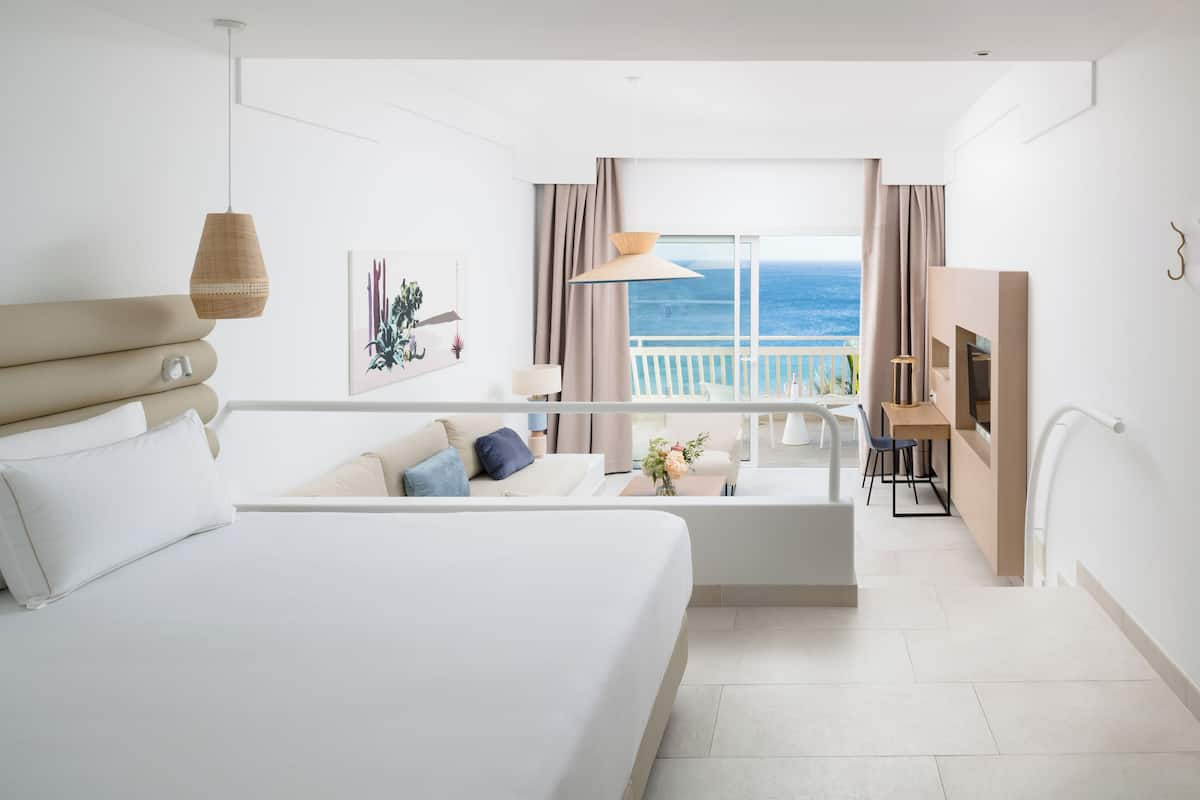 Een moderne hotelkamer met een groot bed, een balkon met uitzicht op zee, een zithoek en een televisie.
