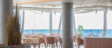 Un restaurante moderno con vista al océano, que cuenta con techos altos, ventanas amplias y mobiliario contemporáneo.