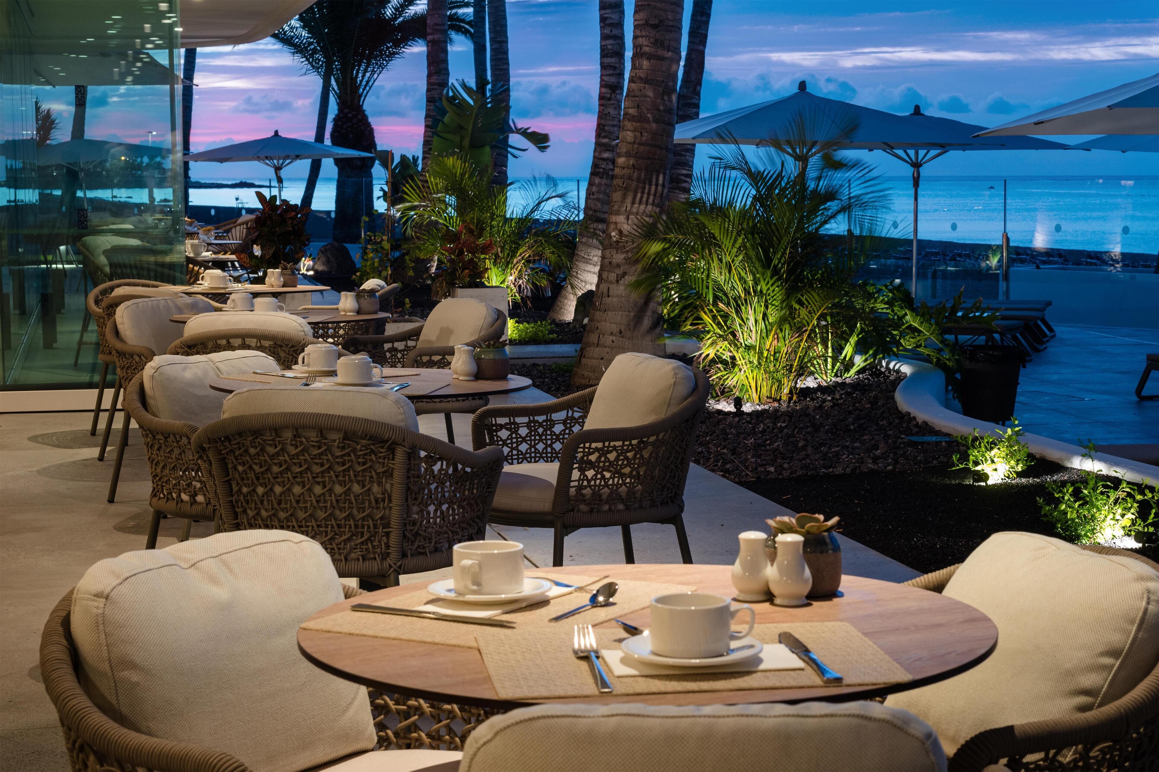 An outdoor dining area with wicker chairs and tables, overlooking a beach and ocean at dusk.