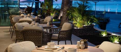 An outdoor dining area with wicker chairs and tables, overlooking a beach and ocean at dusk.