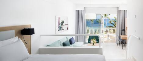 A hotel room with a bed, a desk, a chair, a balcony with a view of the ocean, and a painting on the wall.