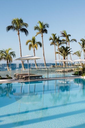 A clear blue pool with lounge chairs and umbrellas, surrounded by palm trees, with a view of the ocean.