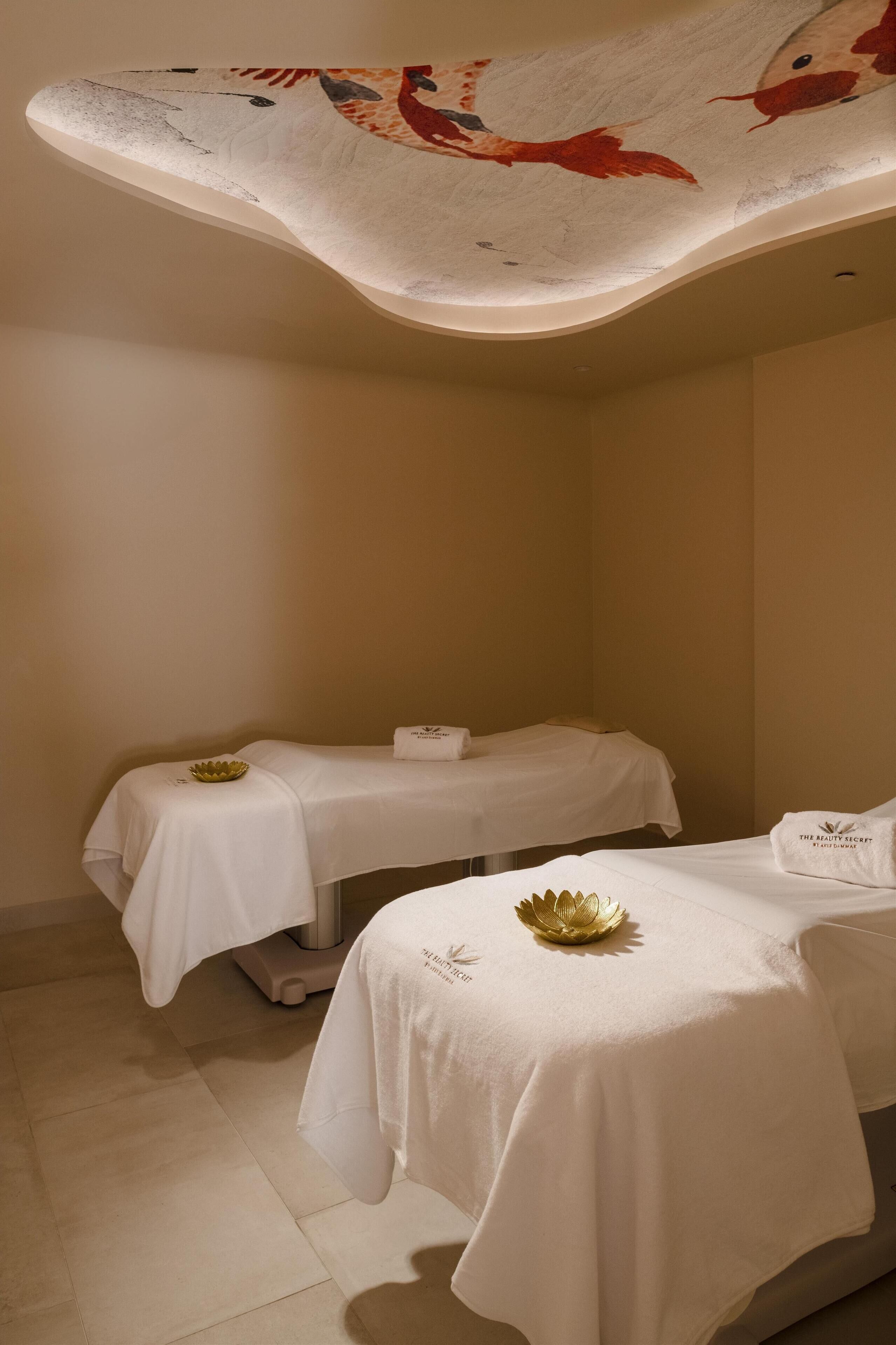 Un cuarto de spa con dos camillas para tratamientos, cada una cubierta con una sábana blanca y un adorno dorado. El techo presenta un mural grande y colorido.