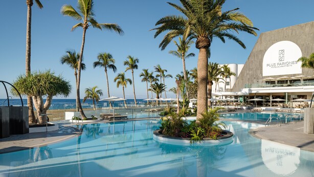 A pool area with palm trees, lounge chairs, and a building with a sign for Plus Fariones Hotels & Apartments. - Plus Fariones Suite Hotel (Tías)