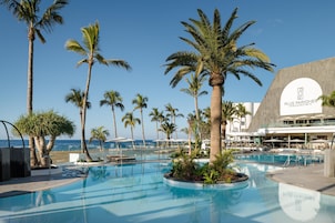 Zona de piscina con palmeras, sillas de descanso y un edificio con un cartel de Plus Fariones Hotels & Apartments.