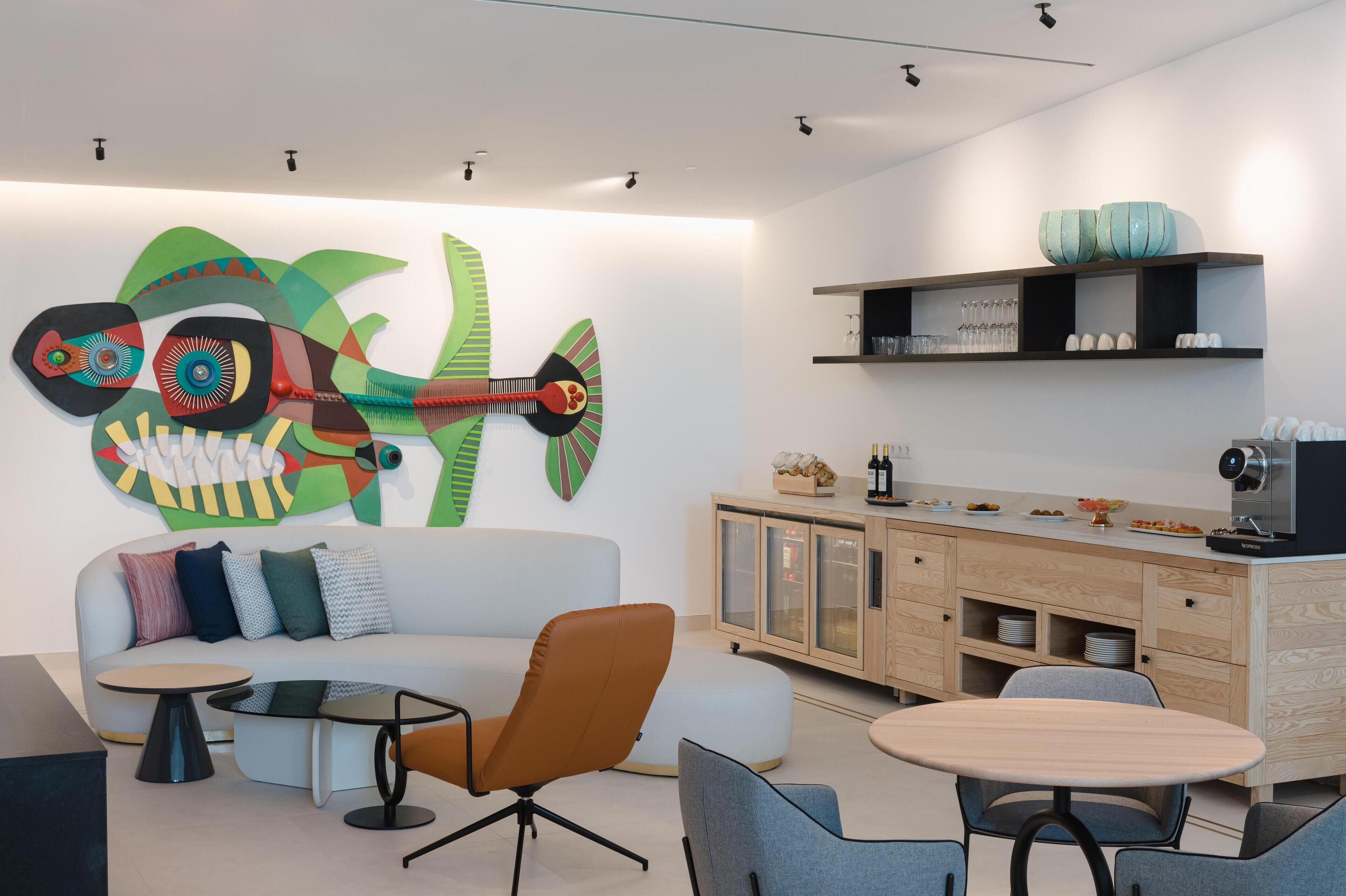 A modern living room with a sofa, a coffee table, and a dining area with chairs. There is a colorful wall art piece and a cabinet with shelves above it.
