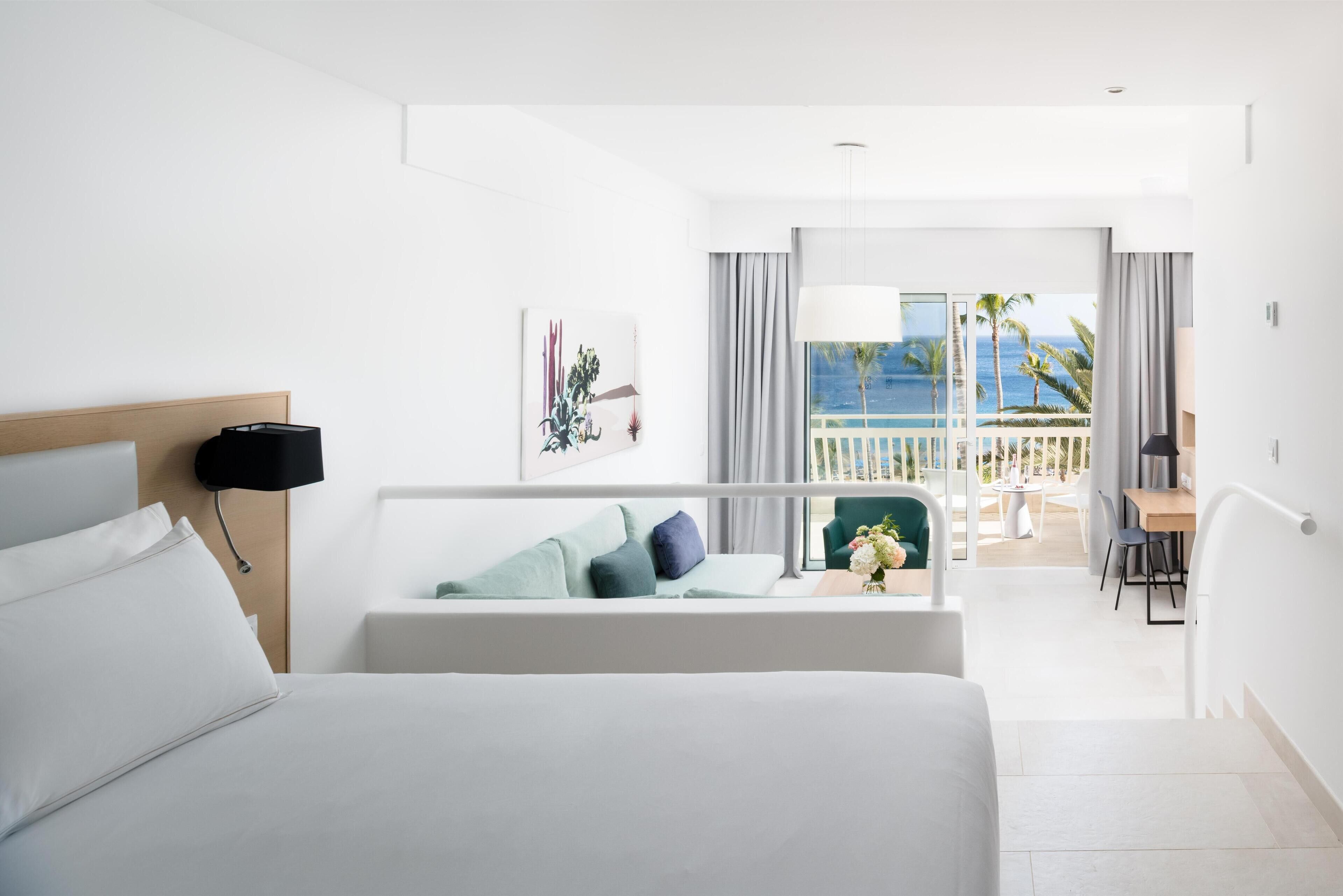 A hotel room with a bed, a desk, a chair, and a balcony with a view of the ocean.