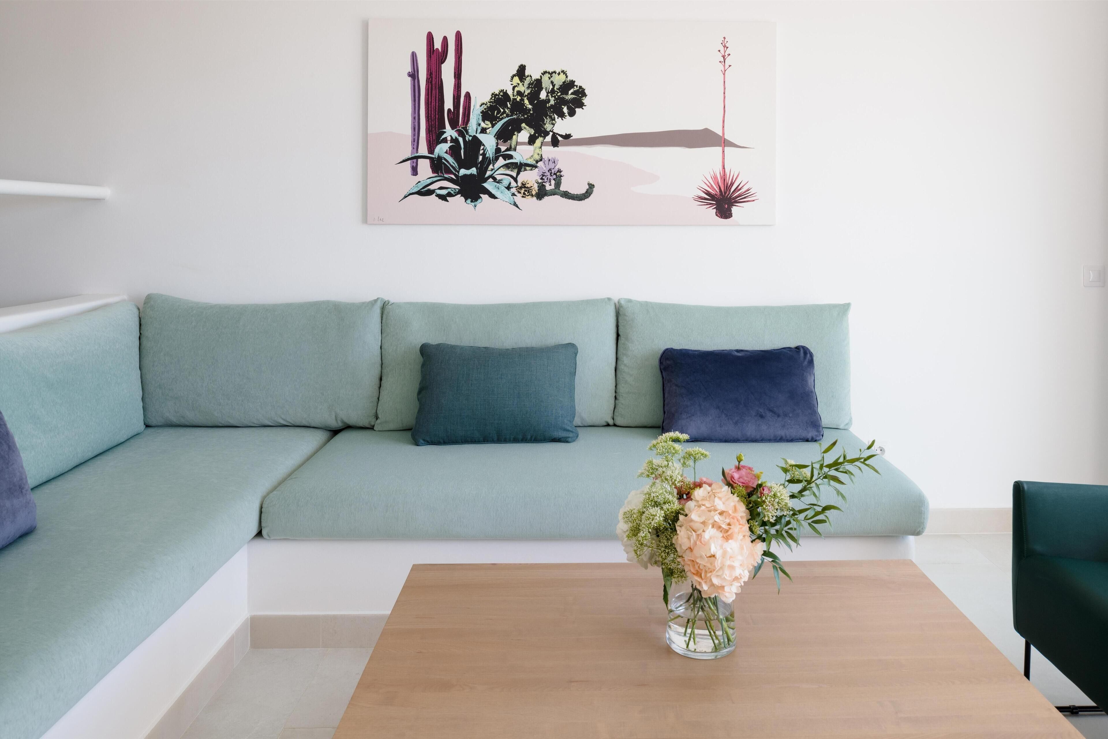 Een moderne woonkamer met een lichtblauwe bank, een houten salontafel met een bloemstuk en een kunstwerk aan de muur met afbeeldingen van cactussen en bloemen.