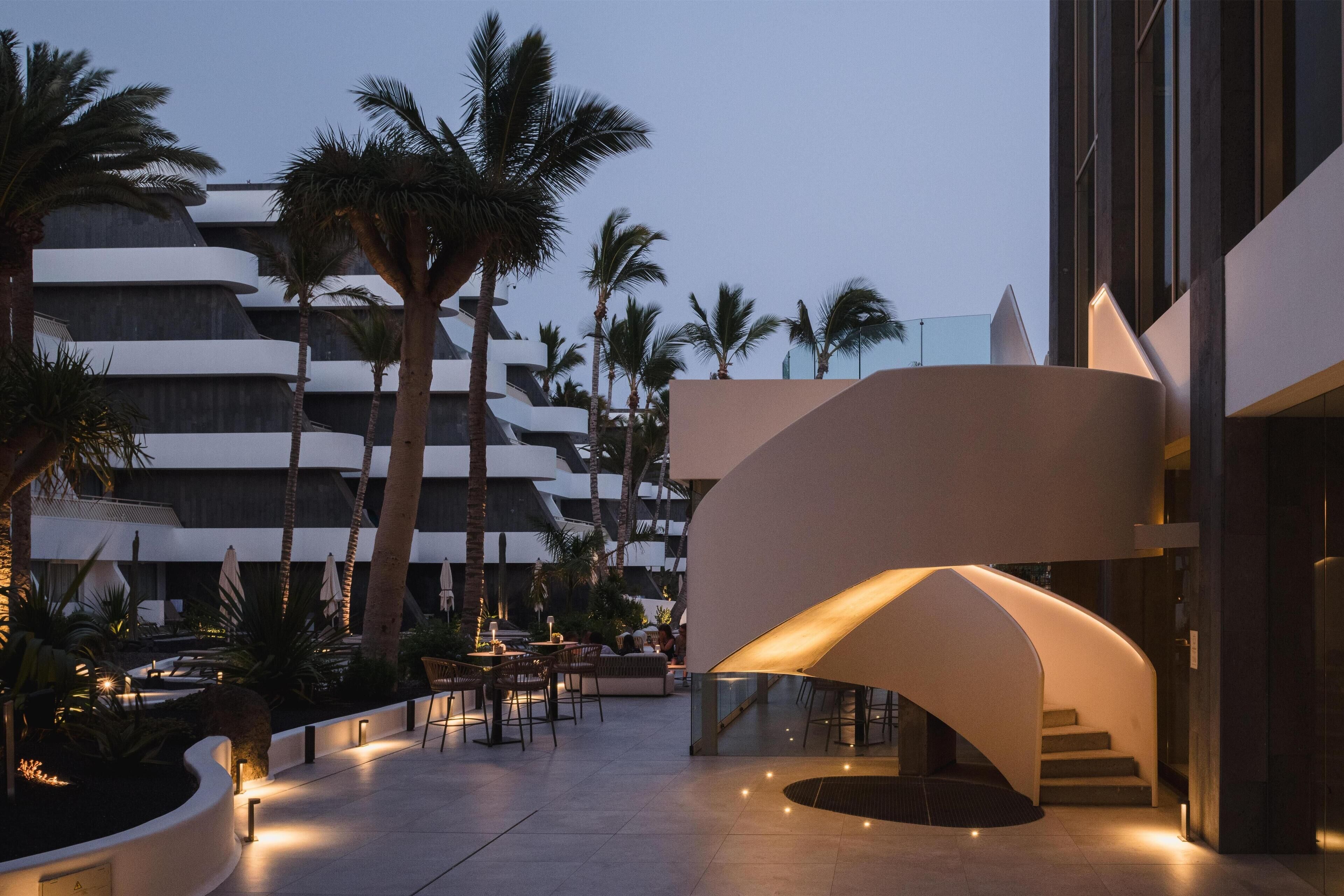 Un hôtel moderne doté d’un escalier à volutes, d’un espace extérieur aménagé avec des sièges, de palmiers et d’une piscine.