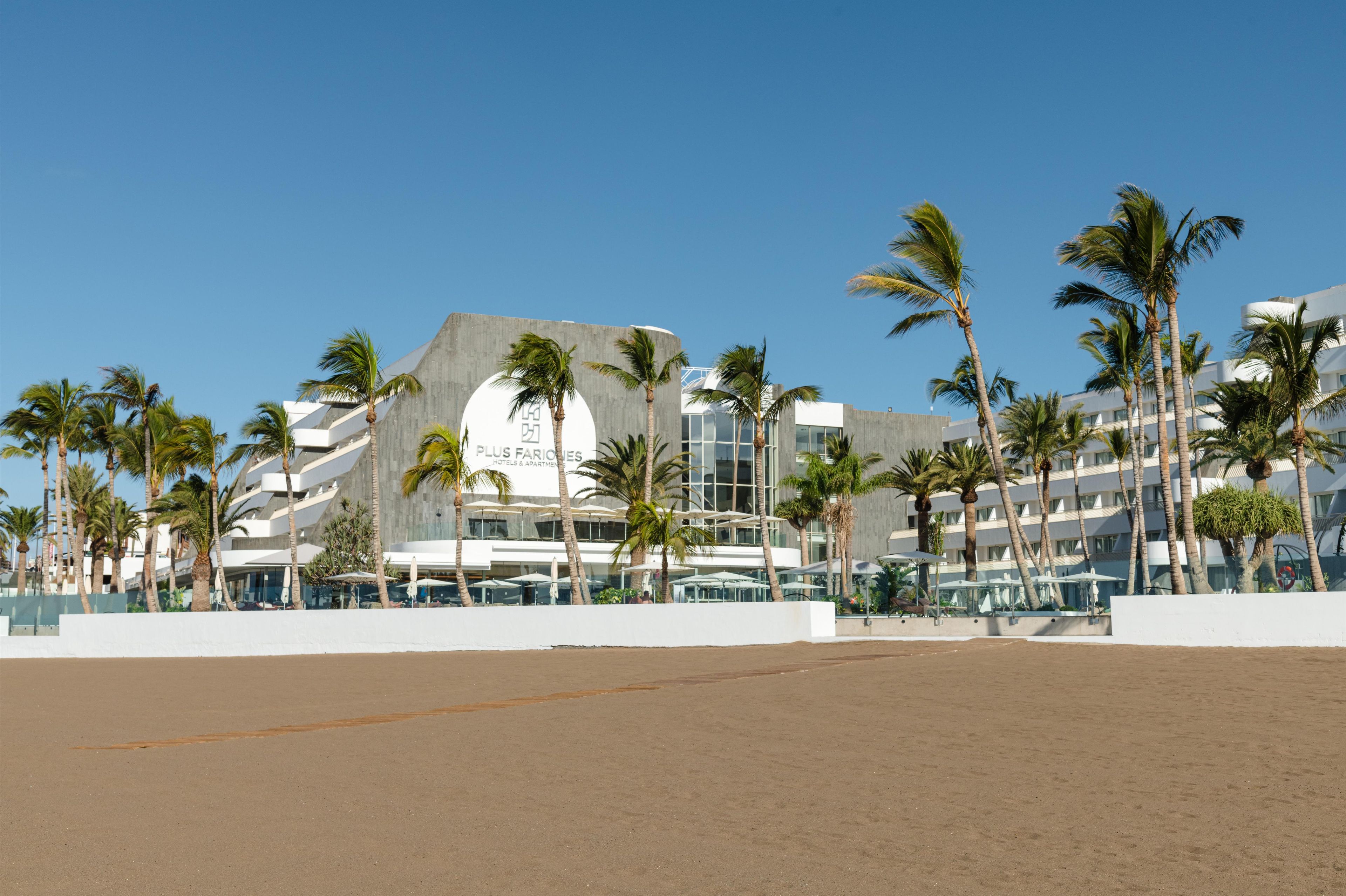 Un hotel moderno con un diseño arquitectónico único, que destaca una estructura circular y varios balcones. Palmeras bordean la playa, y el hotel está junto a una zona de arena.