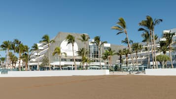 Un hotel moderno con un diseño arquitectónico único, que destaca una estructura circular y varios balcones. Palmeras bordean la playa, y el hotel está junto a una zona de arena.