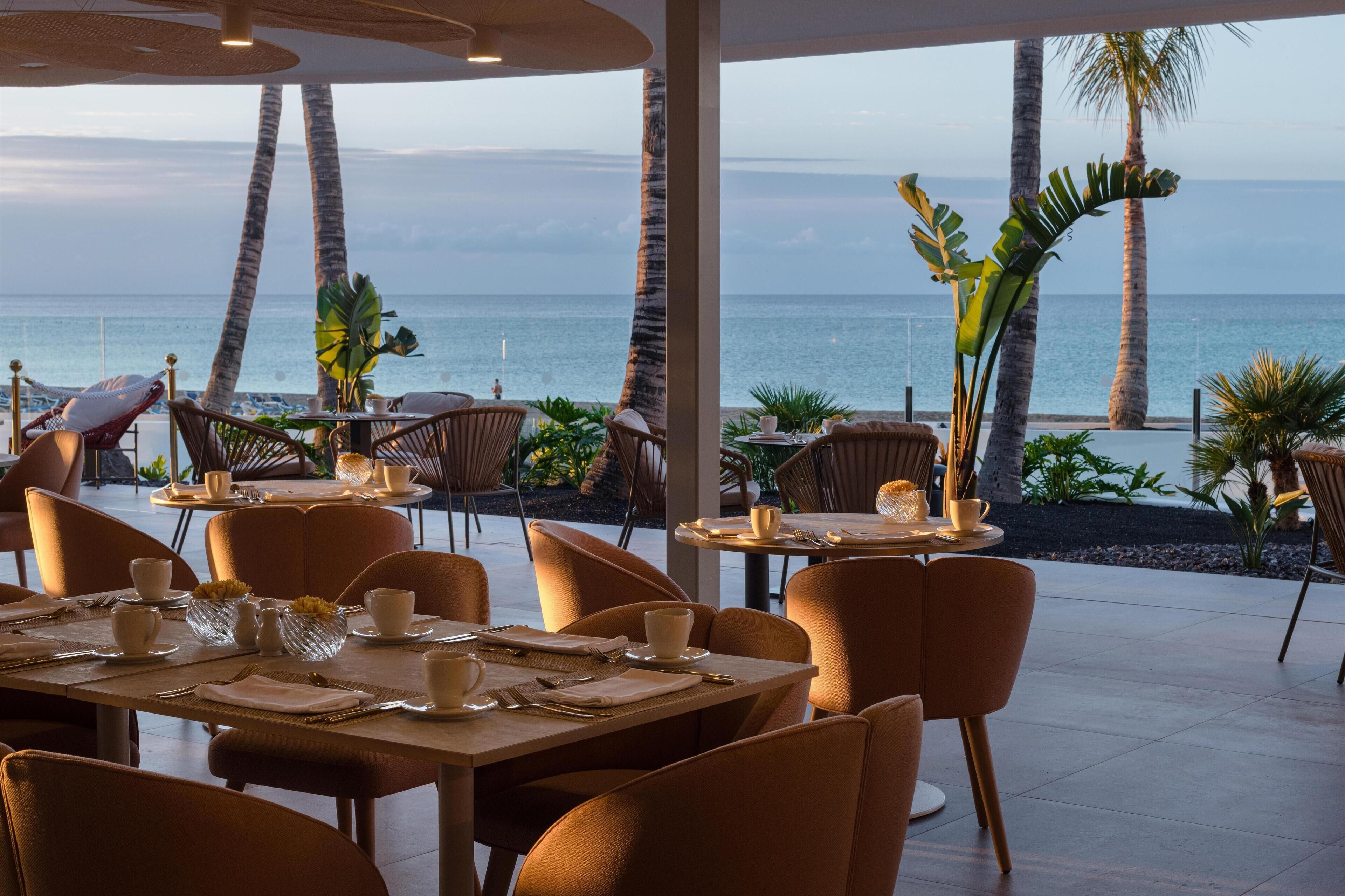 A restaurant with tables and chairs arranged for dining, overlooking a beach with palm trees and the ocean.
