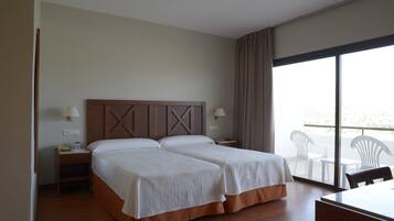 Quarto de hotel com duas camas, cabeceira de madeira, varanda com cadeiras e uma mesa.