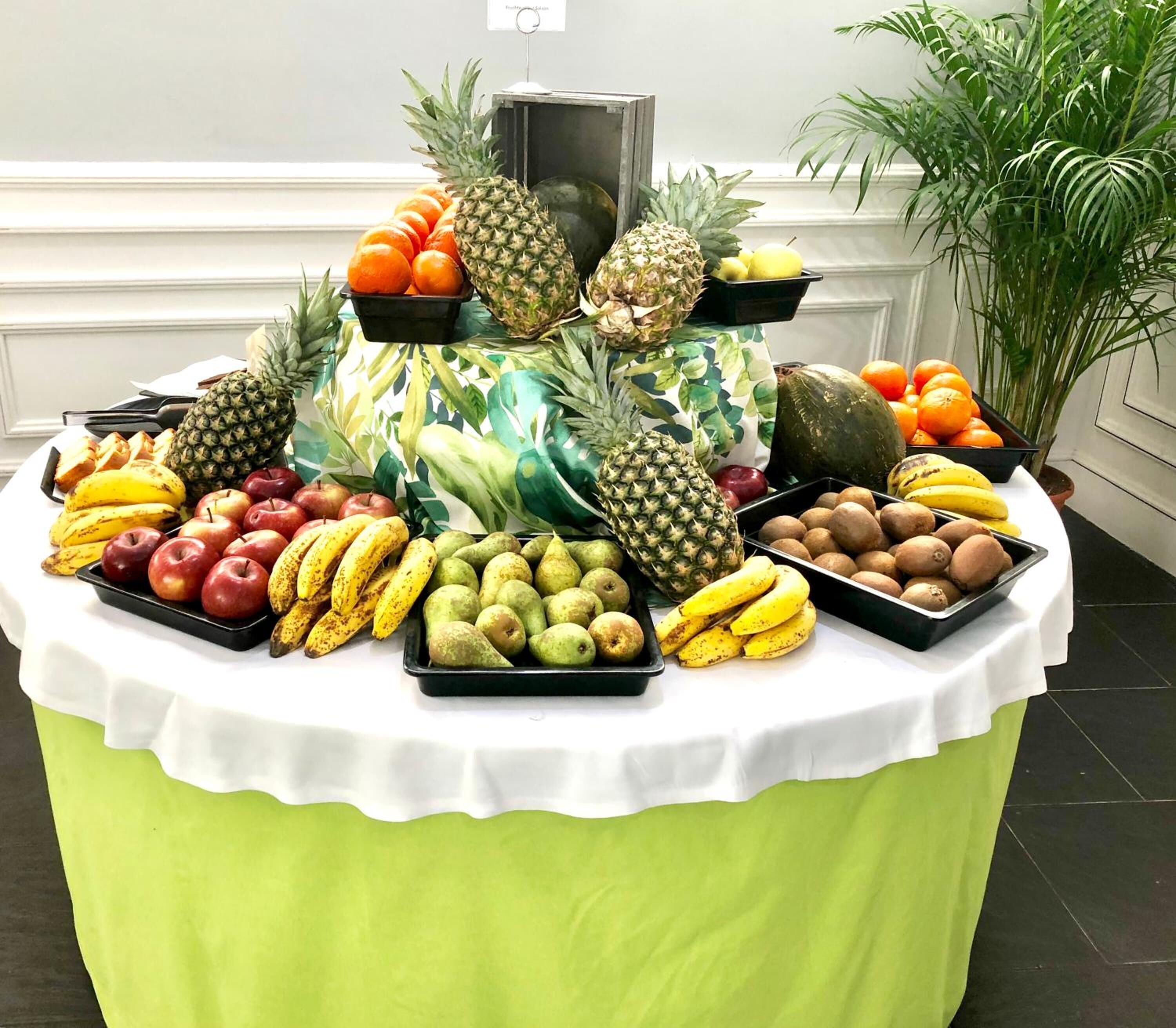 A fruit display table with various fruits and a decorative plant in the background.