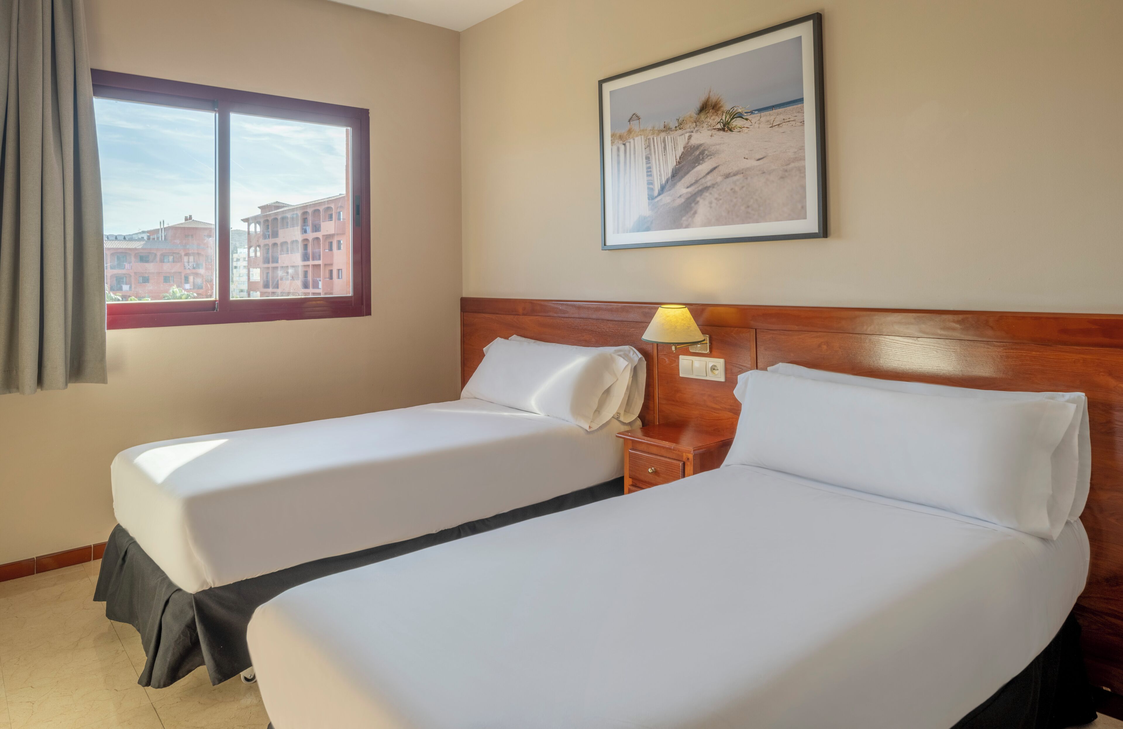 Een hotelkamer met twee bedden, een raam met gordijnen, een lamp en een lijst met een foto aan de muur.