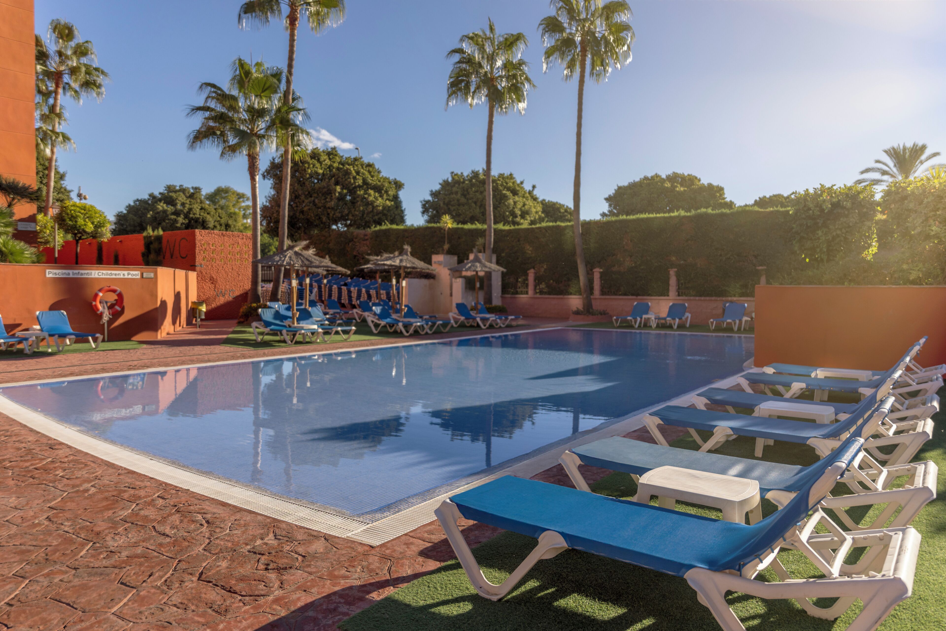 Un espace piscine avec des chaises longues, un espace de jeux pour enfants et des palmiers.