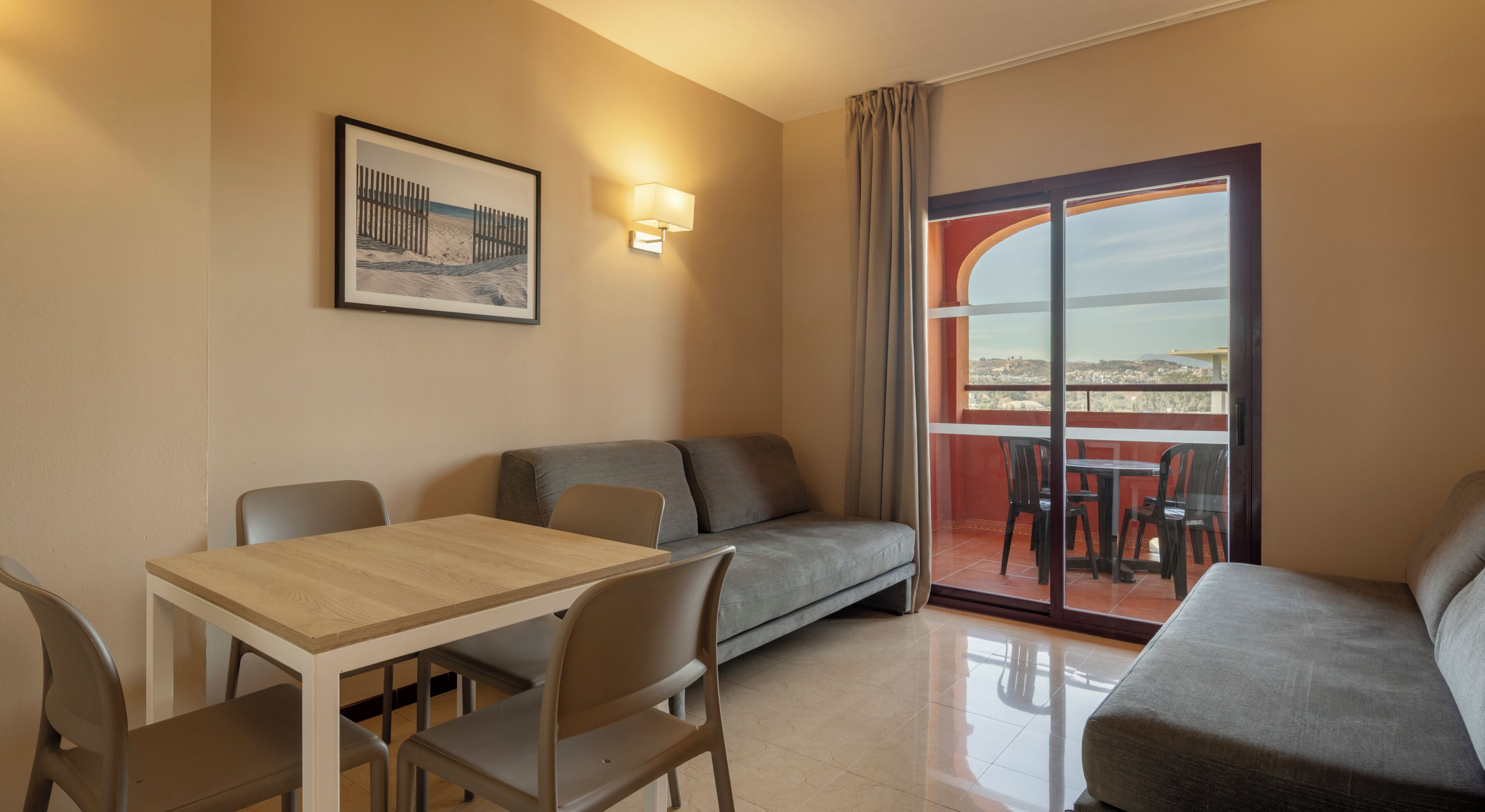 A room with a dining table, two sofas, a balcony with a view, and a framed picture on the wall.