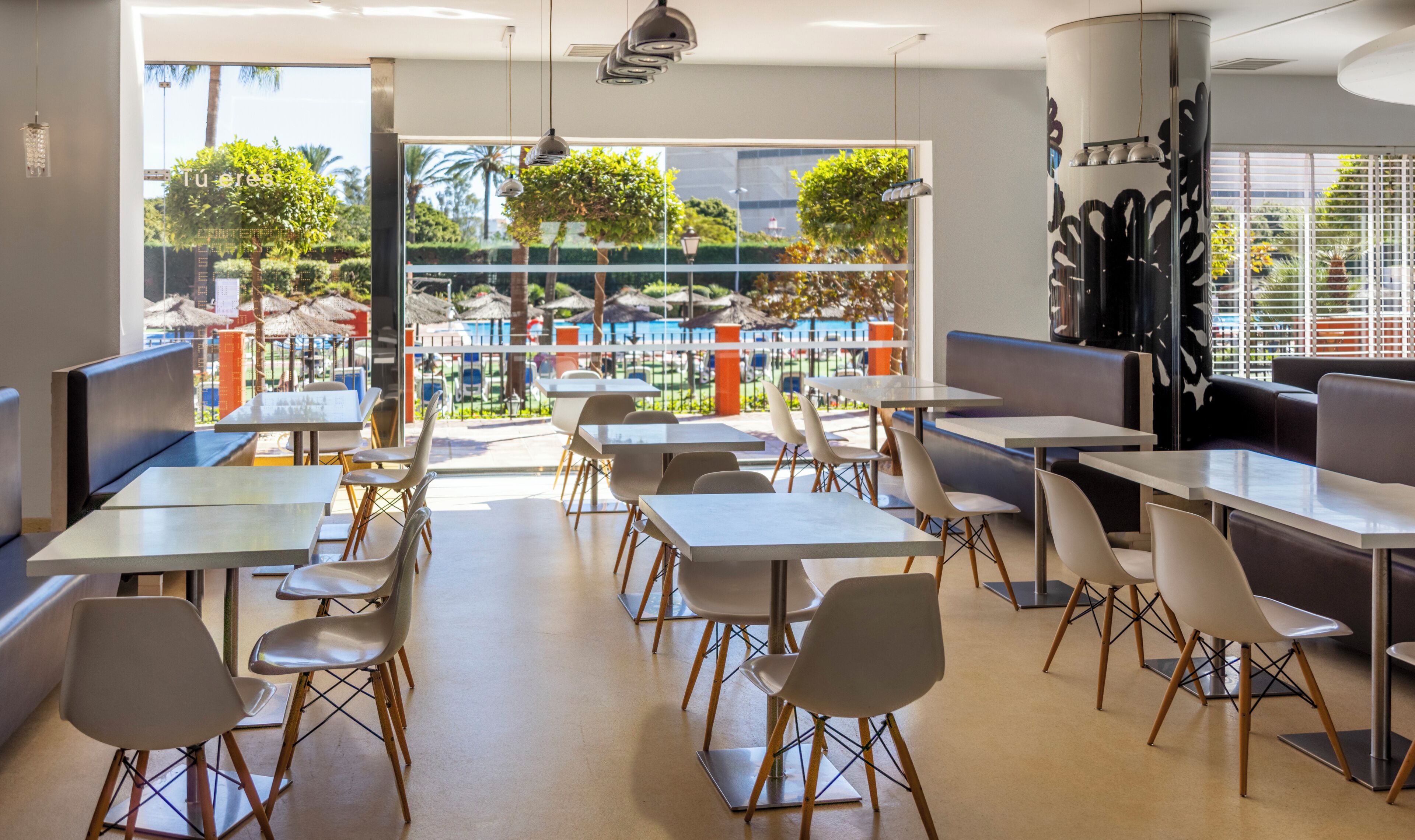 Een modern restaurantinterieur met tafels en stoelen, grote ramen met uitzicht op een zwembad en groen.
