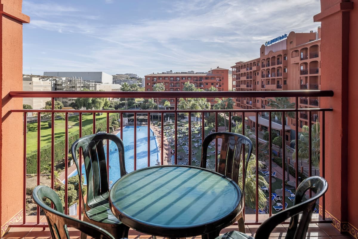 Un balcón con mesa y sillas, con vista a una piscina y el edificio de un hotel.