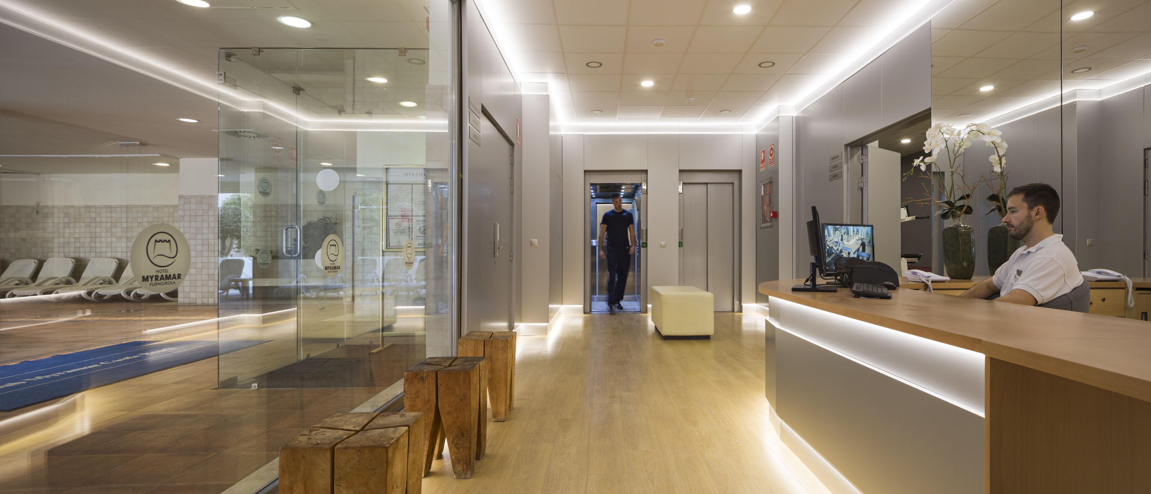 A hotel reception area with a wooden reception desk, a computer, and a large mirror. There are wooden stools and a glass wall with a view of the lobby.