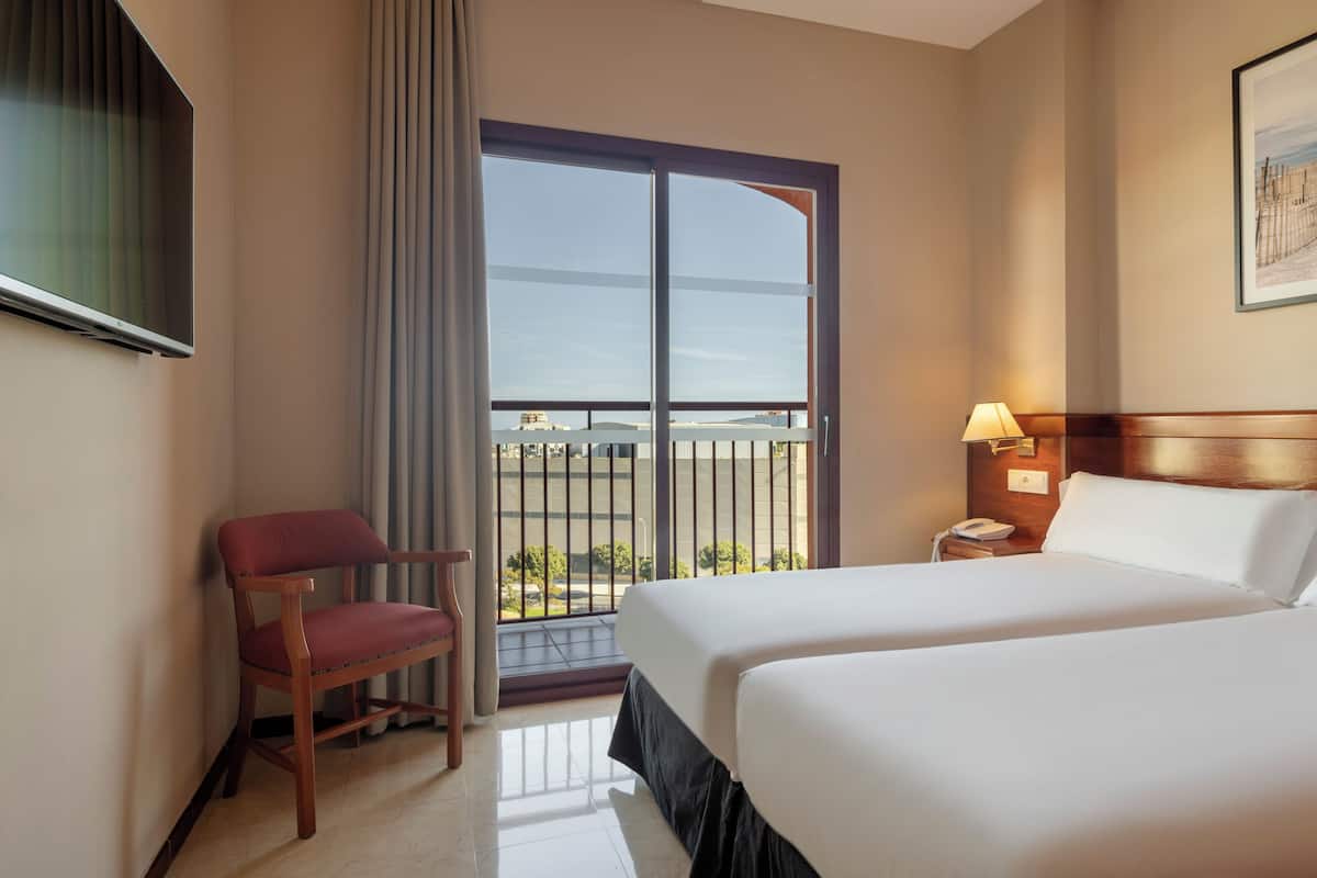 Habitación de hotel con una cama grande, una silla, un escritorio con lámpara, teléfono y ventana con vistas al exterior.