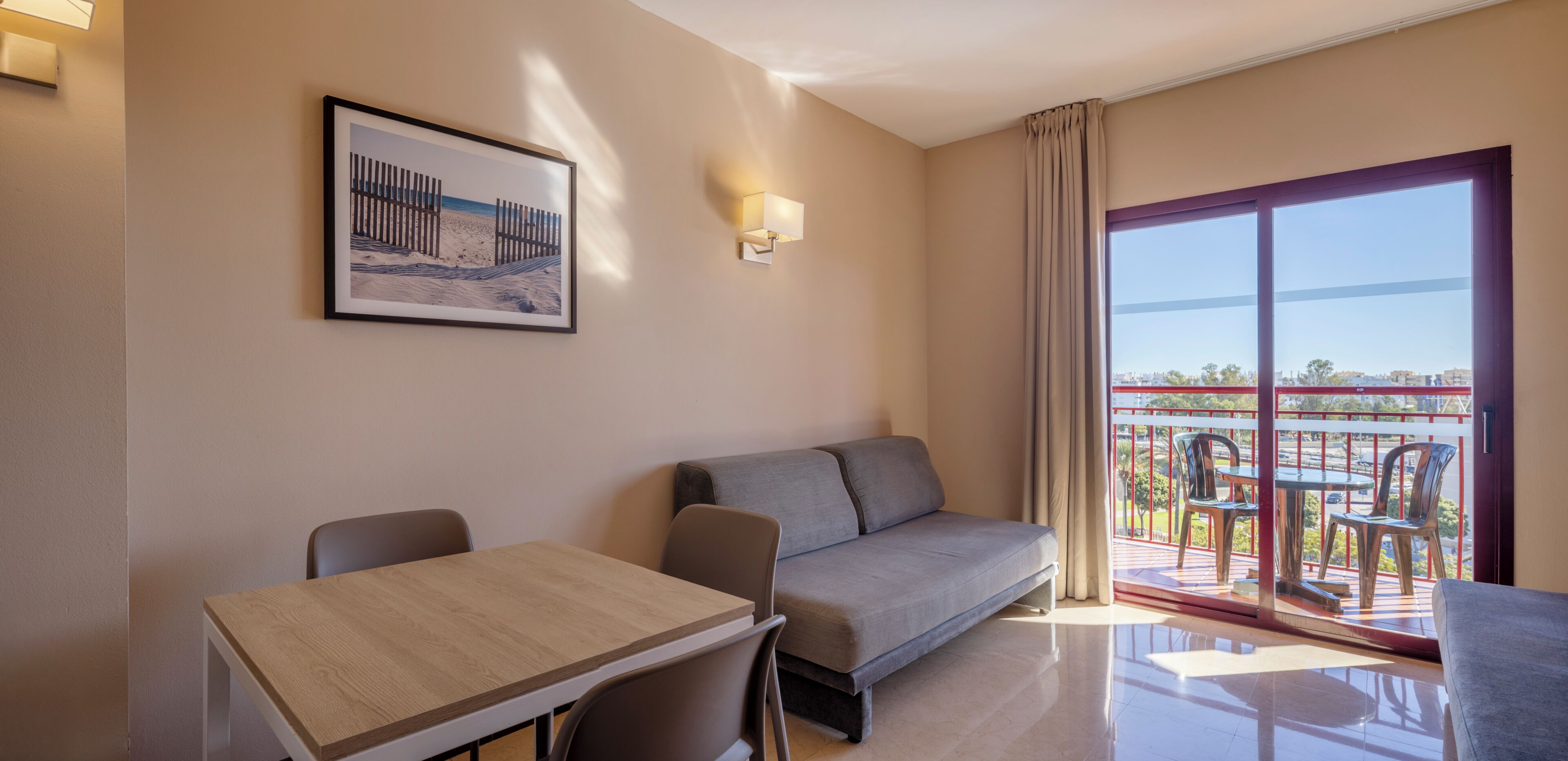 Een hotelkamer met een bank, een eettafel, een balkon met stoelen en een ingelijst schilderij aan de muur.