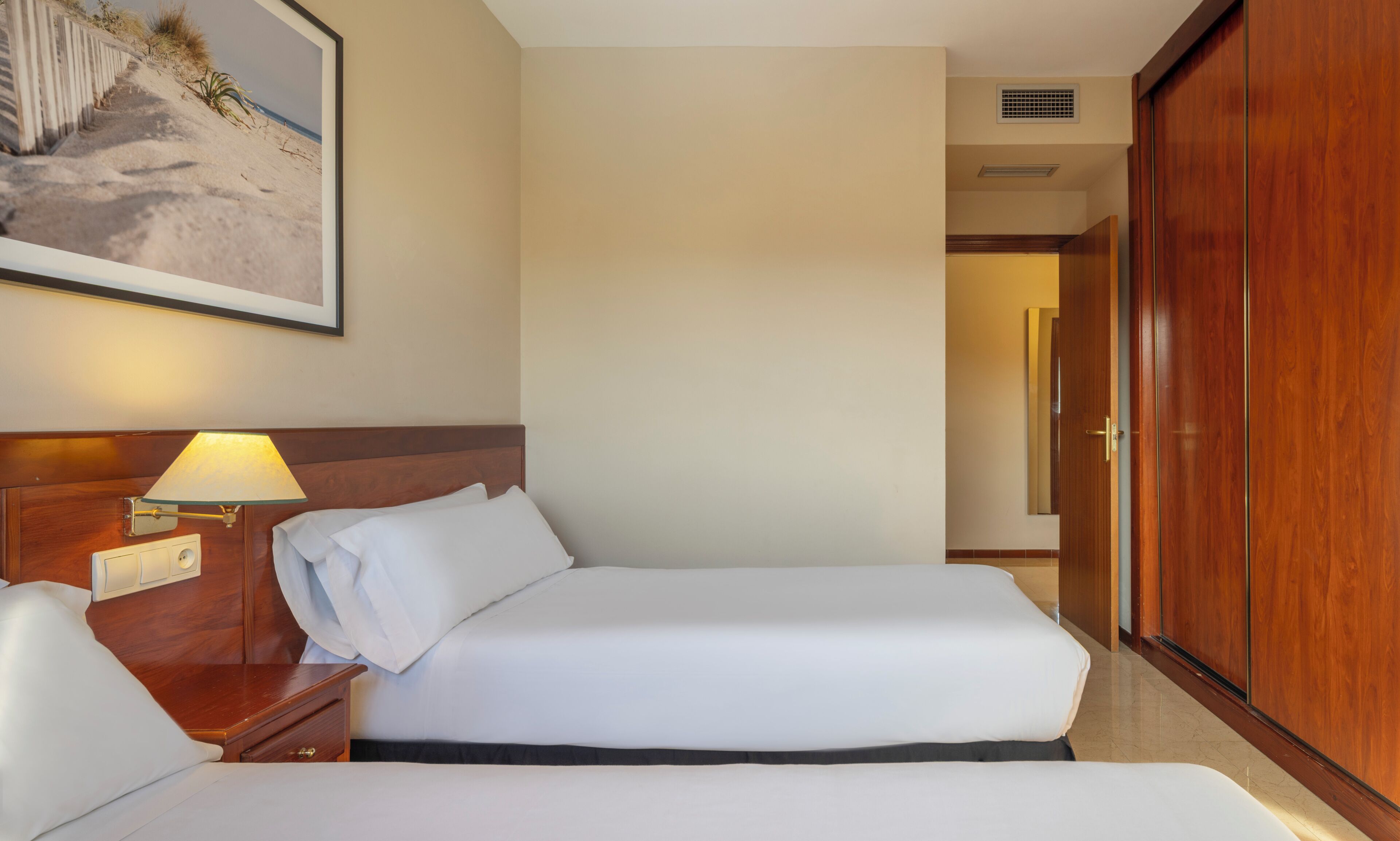 A hotel room with a bed, bedside table, lamp, and a large framed picture of a beach.