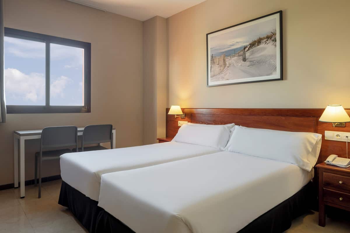 Habitación de hotel con dos camas, un escritorio, sillas, una lámpara, un teléfono y un cuadro en la pared.