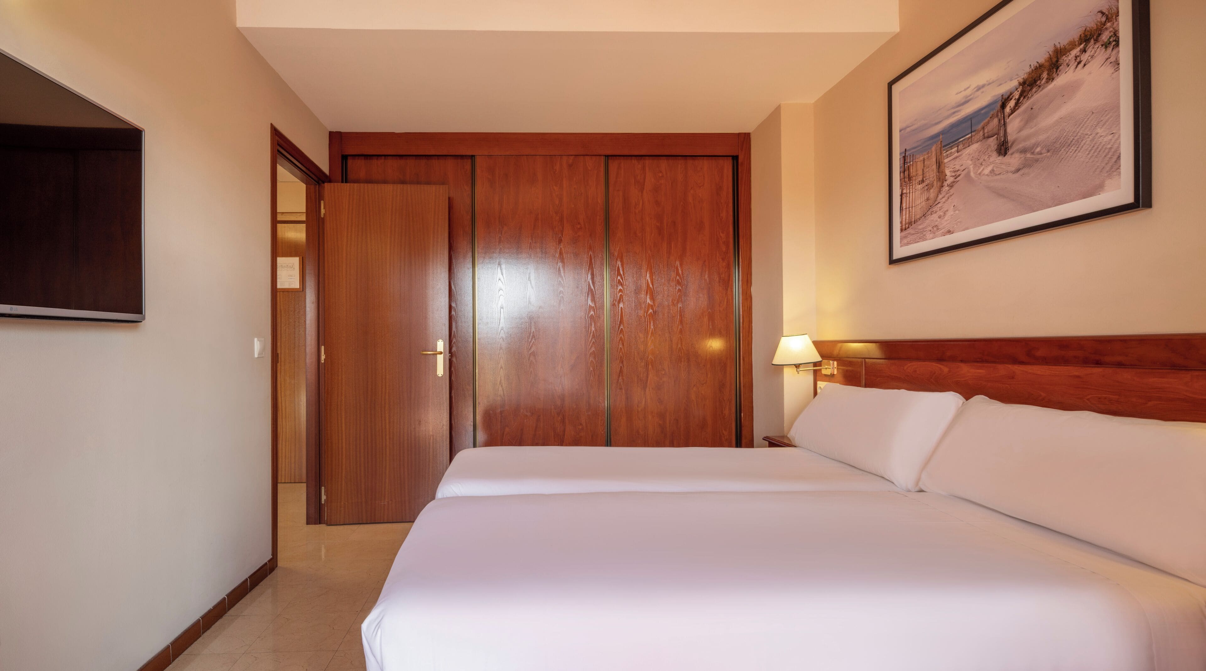 Habitación de hotel con un armario de madera, una cama con ropa de cama blanca, una mesita de noche con lámpara y un cuadro enmarcado en la pared.