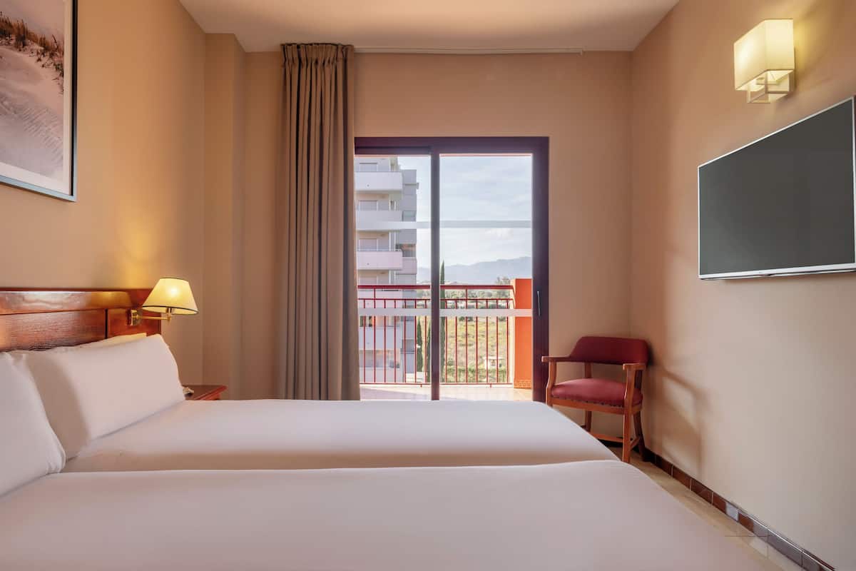 Habitación de hotel con cama, una silla, televisor, un cuadro y balcón con vistas a edificios.