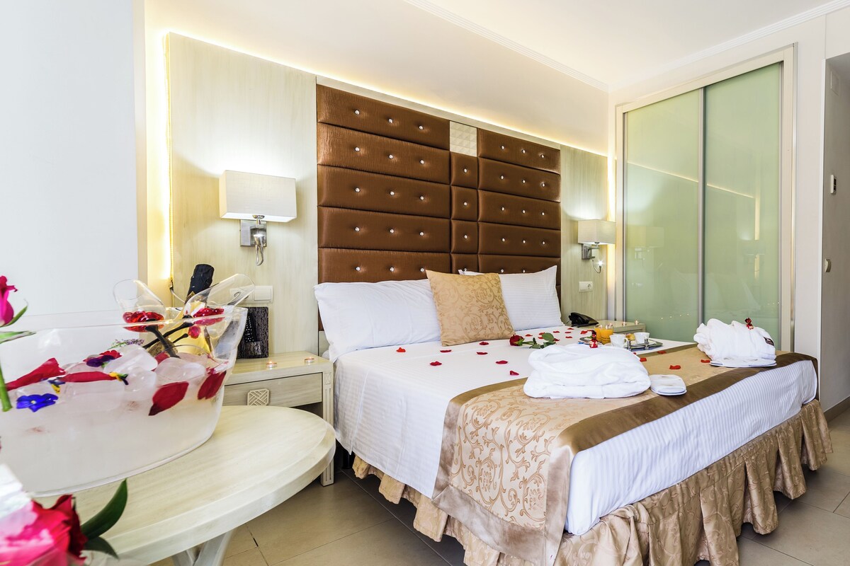 A hotel room with a large bed, a bedside table with a lamp, a glass-enclosed wardrobe, and a small round table with a floral arrangement.