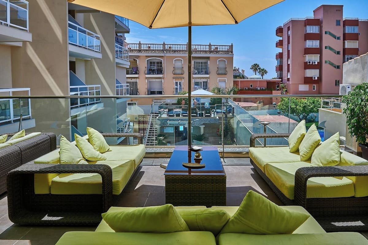 Terraza con mobiliario de mimbre y cojines amarillos, con vistas a una piscina y otros edificios.