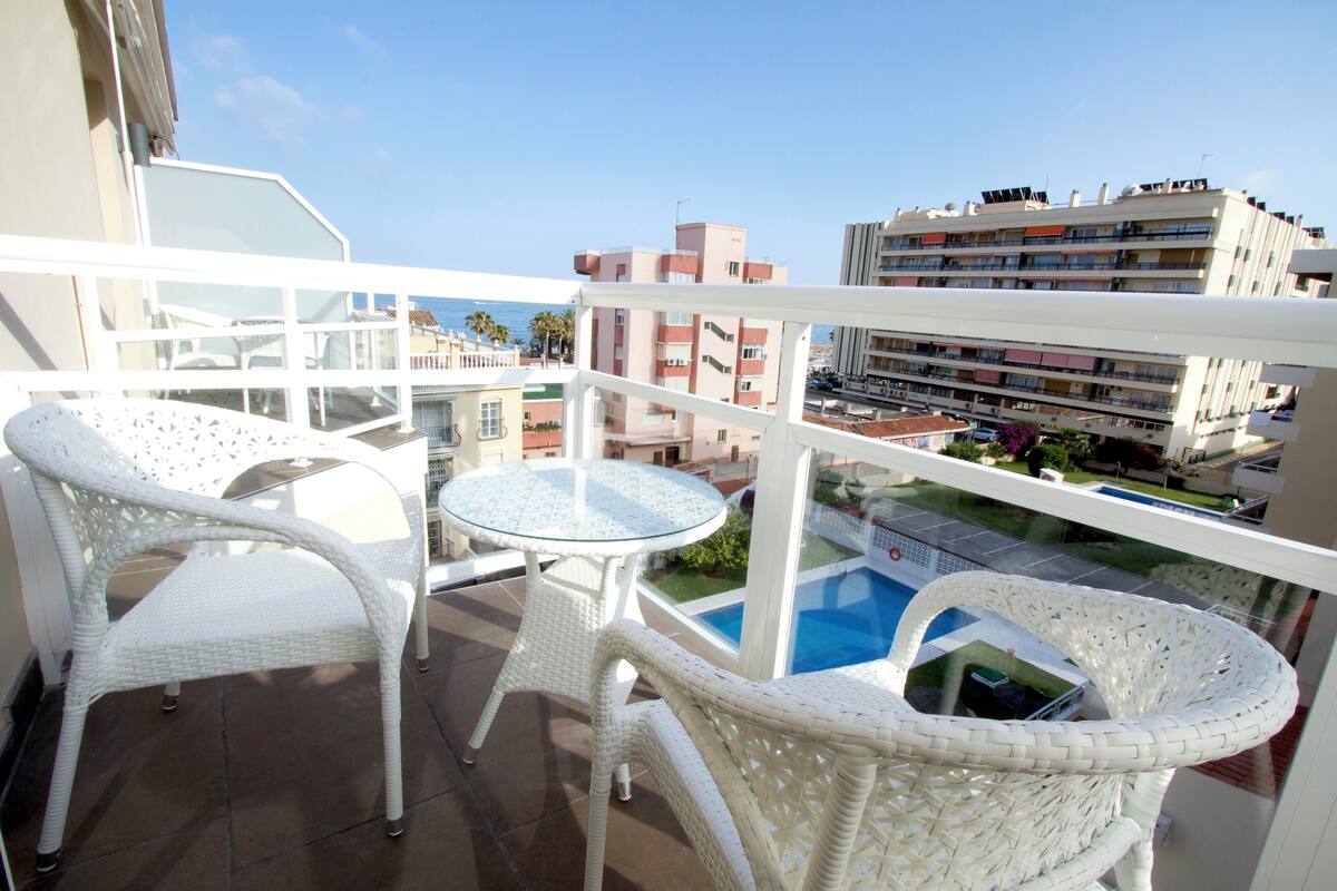 Un balcón con mobiliario de mimbre blanco, una mesa de vidrio y vistas a una piscina y edificios.