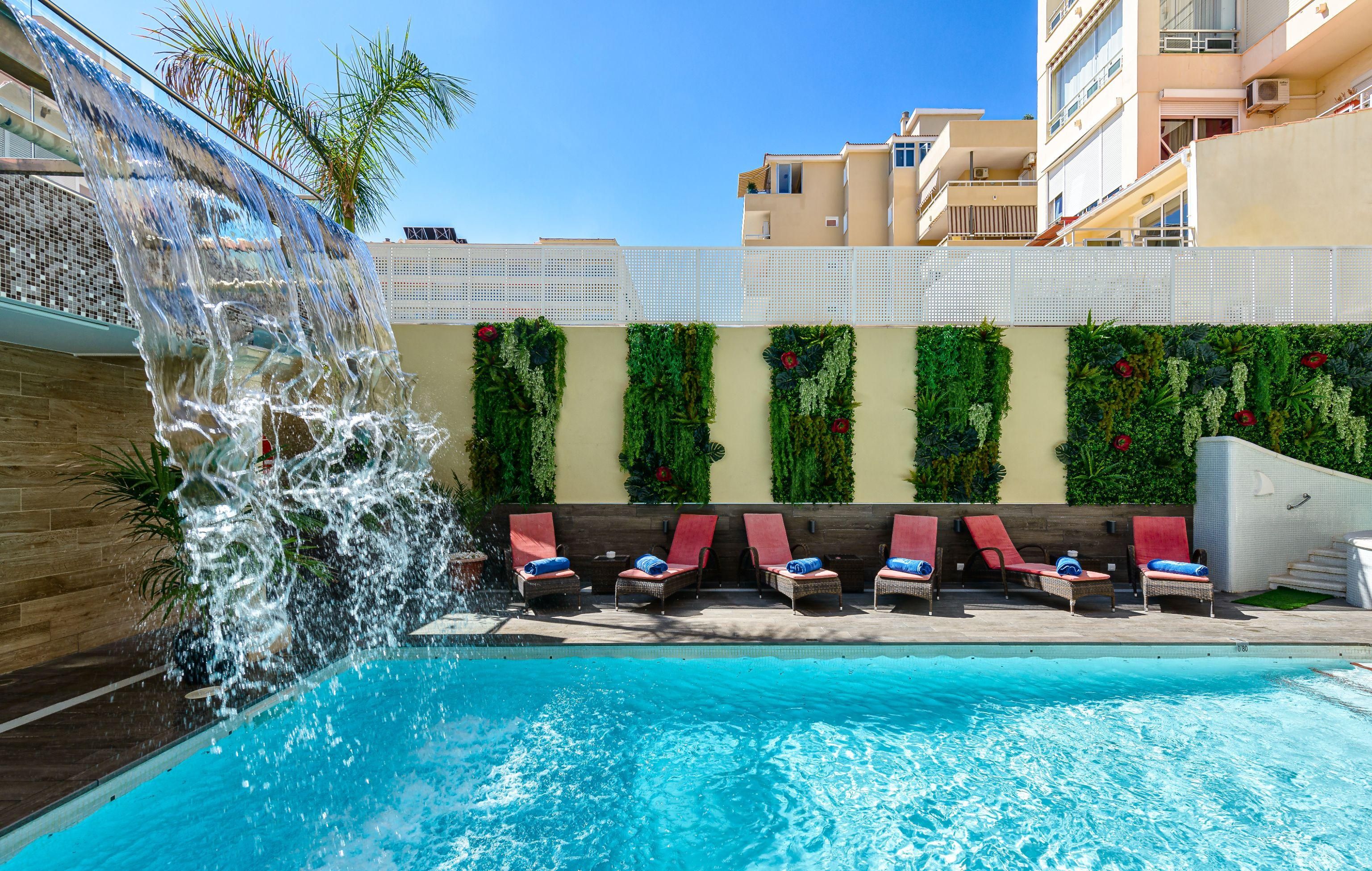 A swimming pool with a waterfall feature, surrounded by lounge chairs and a wall decorated with greenery.