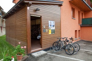 Un edificio de madera con una puerta abierta, mostrando bicicletas en su interior. Hay carteles en la pared y un aparcamiento para bicicletas afuera.