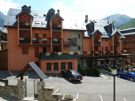 Un hotel de varios pisos con balcones decorados con flores, un estacionamiento y un paisaje montañoso al fondo.