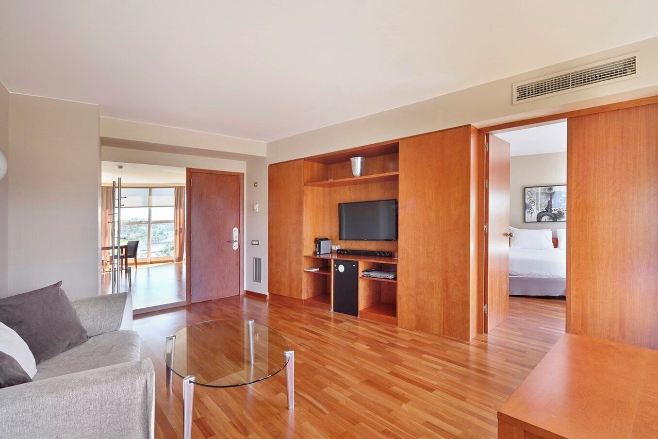 A modern living room with a glass coffee table, a sofa, a flat-screen TV, and a kitchen area with wooden cabinets.