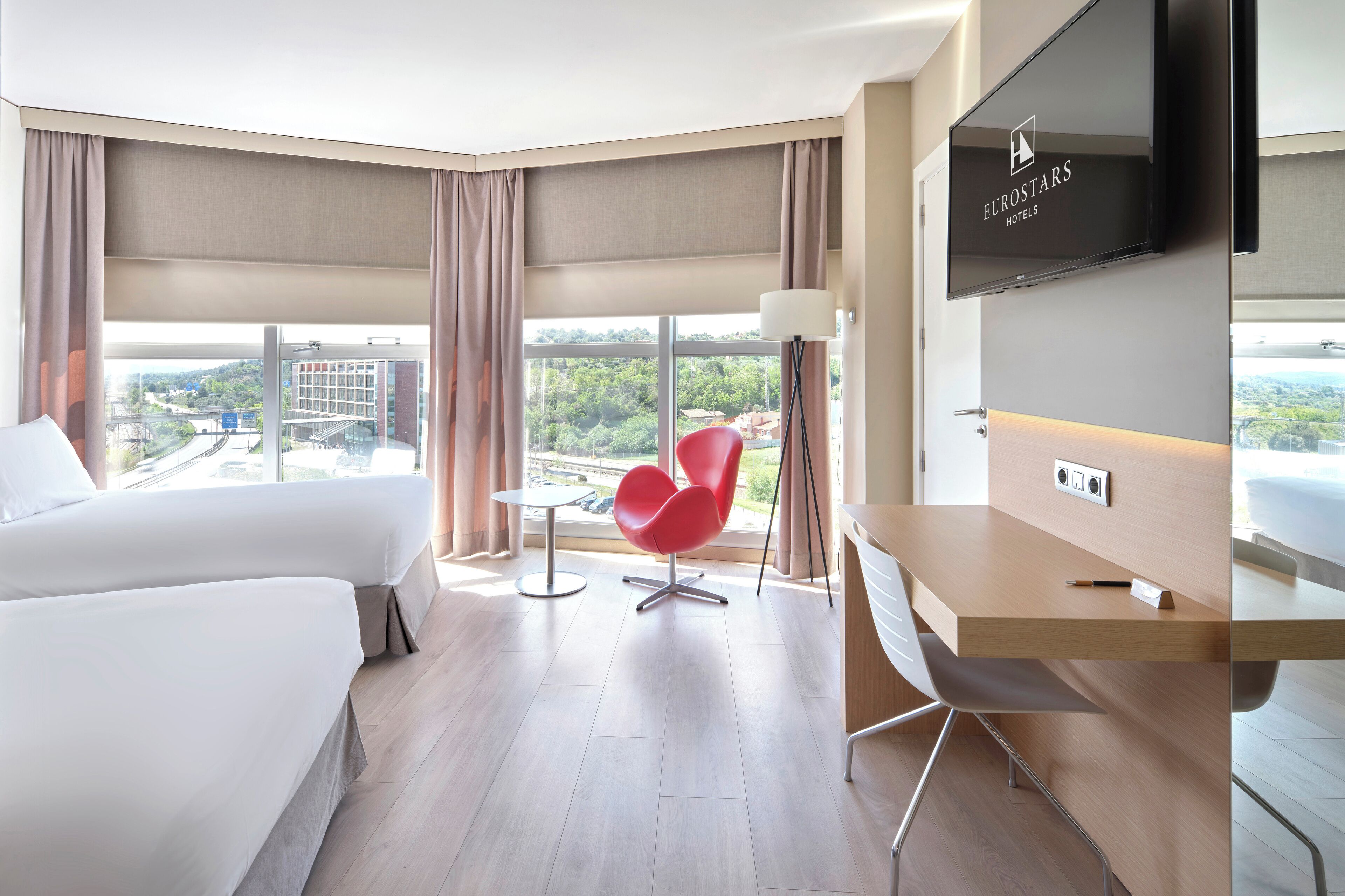 A modern hotel room with a large bed, a desk, a red chair, and a TV.