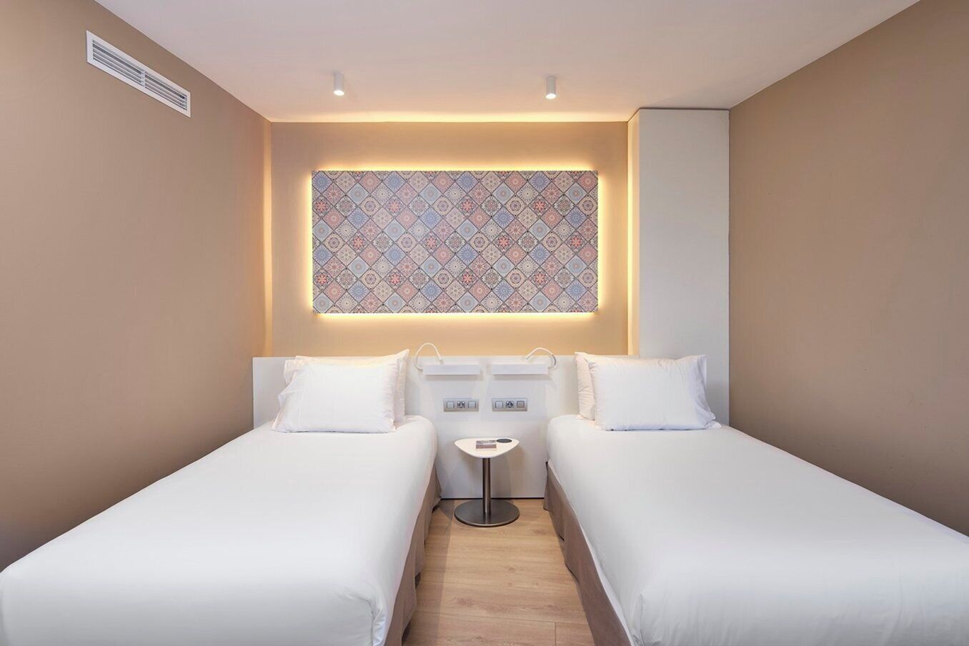 A hotel room with two single beds, a small bedside table, a wall-mounted air vent, and a patterned window.