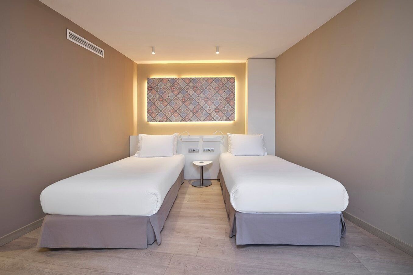A hotel room with two single beds, a small bedside table, a wall-mounted air vent, and a patterned wall decoration.