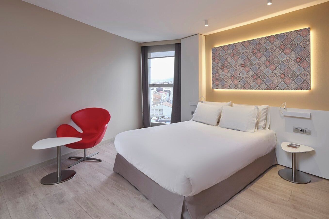 Una habitación de hotel moderna con una cama grande, una silla roja, una mesita y un adorno de pared con diseño.