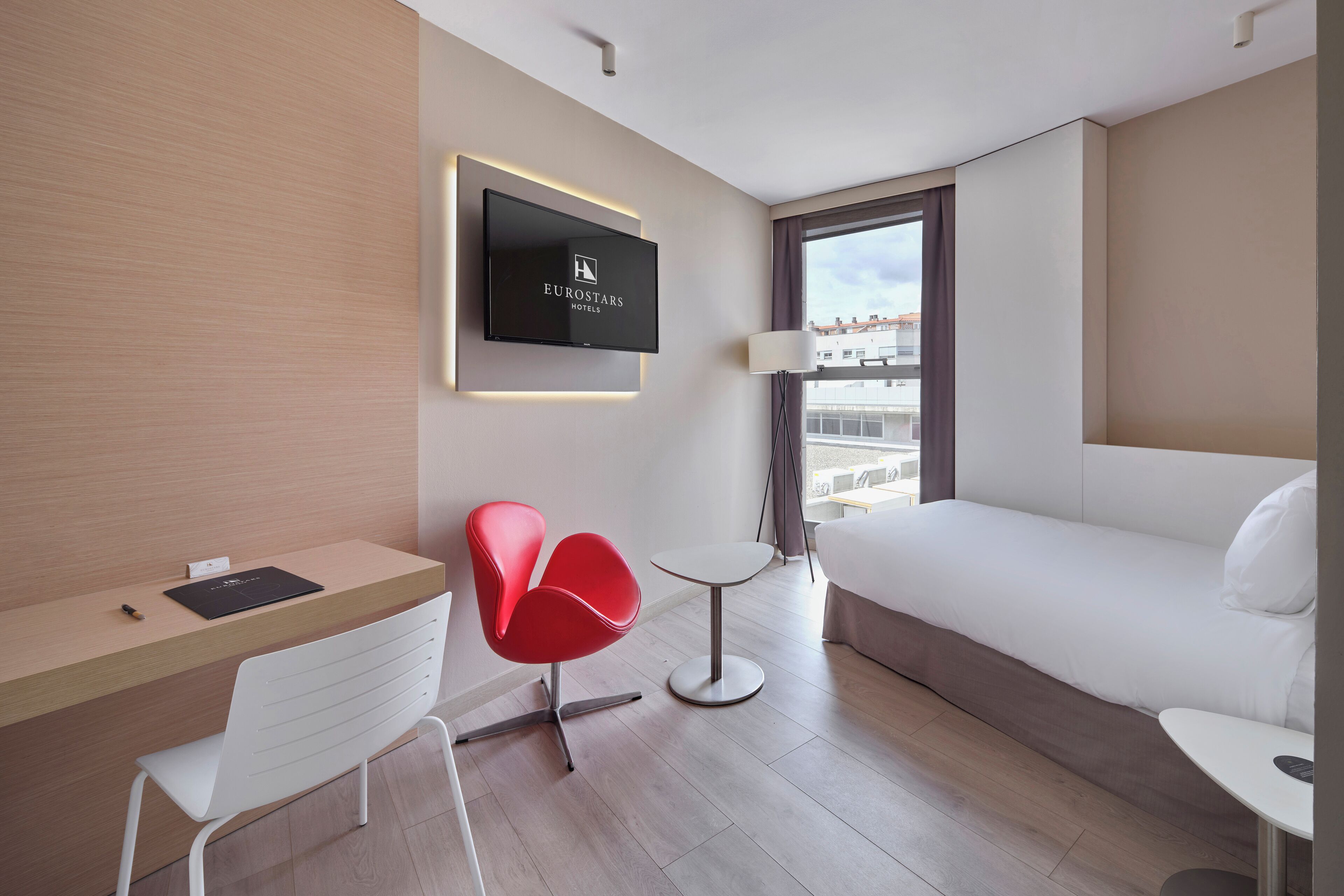 Una habitación de hotel moderna con televisión de pantalla plana, un escritorio con silla, una mesita redonda pequeña, un sofá y una ventana con vistas a edificios.
