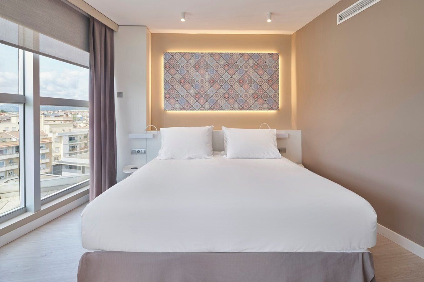 A modern hotel room with a large bed, a bedside table, a nightstand, and a view of the city through large windows.