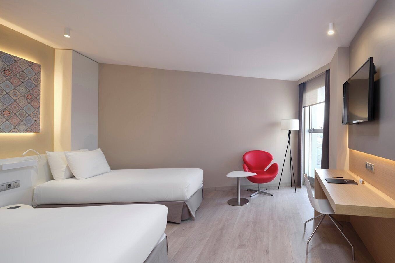 A hotel room with two beds, a red chair, a desk, a flat-screen TV, and a patterned wall.