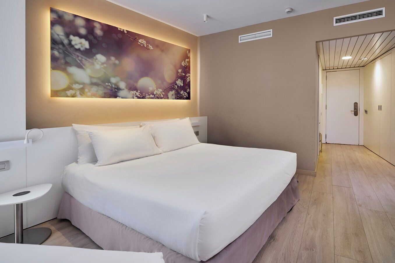Ein Hotelzimmer mit einem großen Bett, einem Nachttisch, einem Wandbild und einer Tür zu einem anderen Raum.