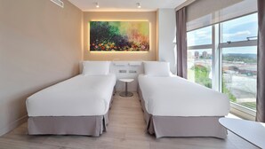 Una habitación de hotel moderna con dos camas individuales, una mesita redonda pequeña, un ventanal grande con vista a la ciudad y un cuadro abstracto colorido en la pared.