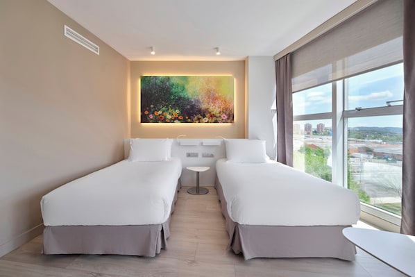 Une chambre d’hôtel moderne avec deux lits simples, une petite table ronde, une grande fenêtre donnant sur la ville et une peinture abstraite aux couleurs vives accrochée au mur.