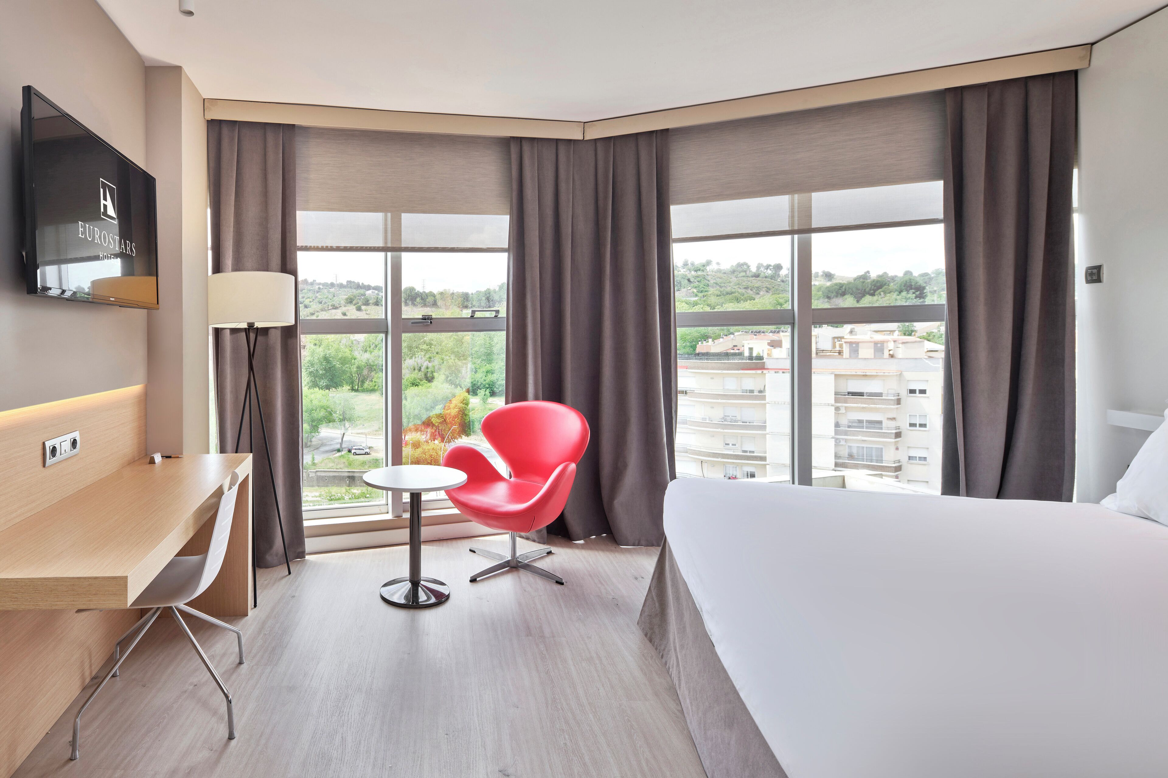 Habitación de hotel con cama, escritorio, silla, televisión y ventanas amplias con cortinas.